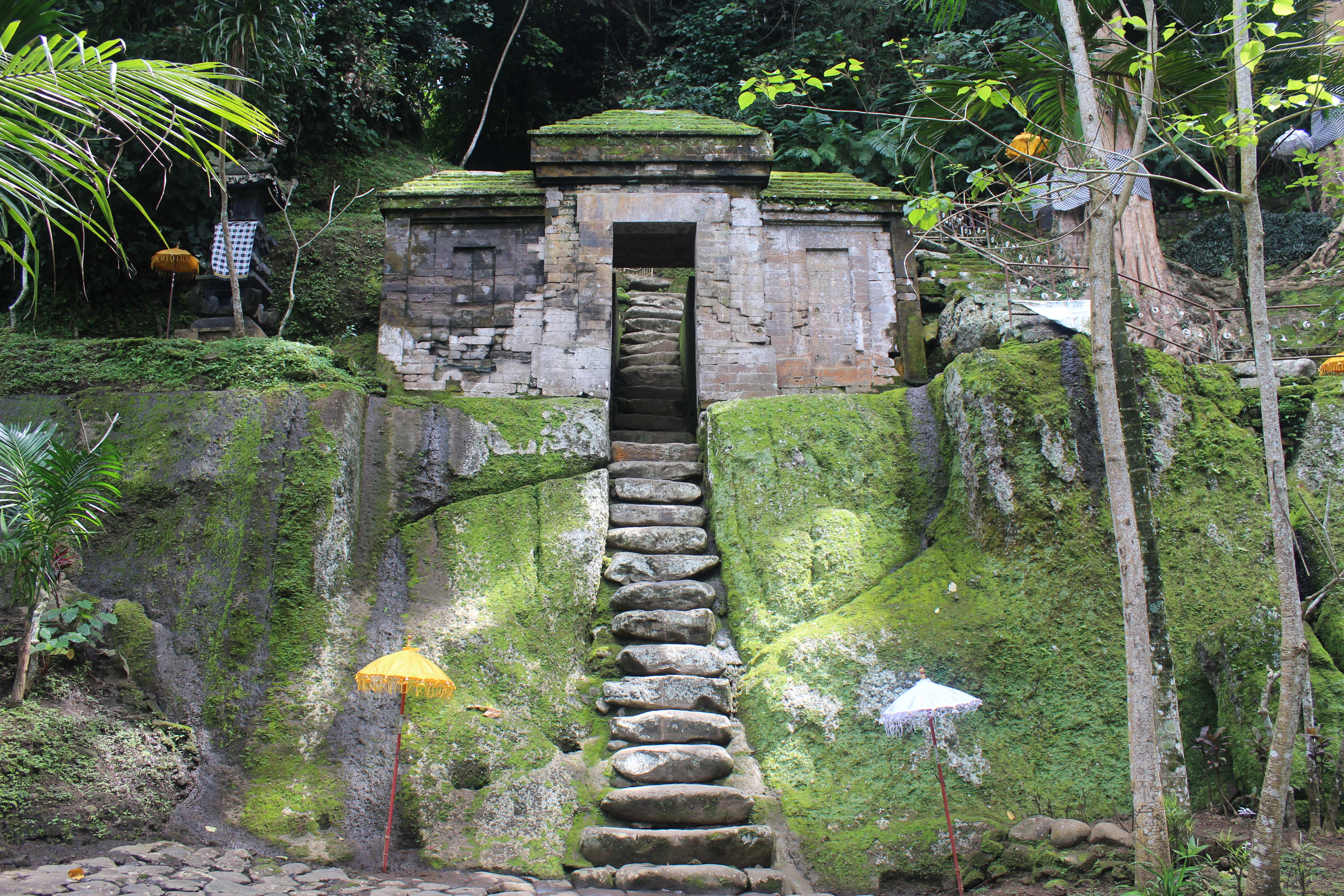 In the jungle, a flight of rock-cut stairs leading up to a gate and shrine complex, with yellow and white umbrellas