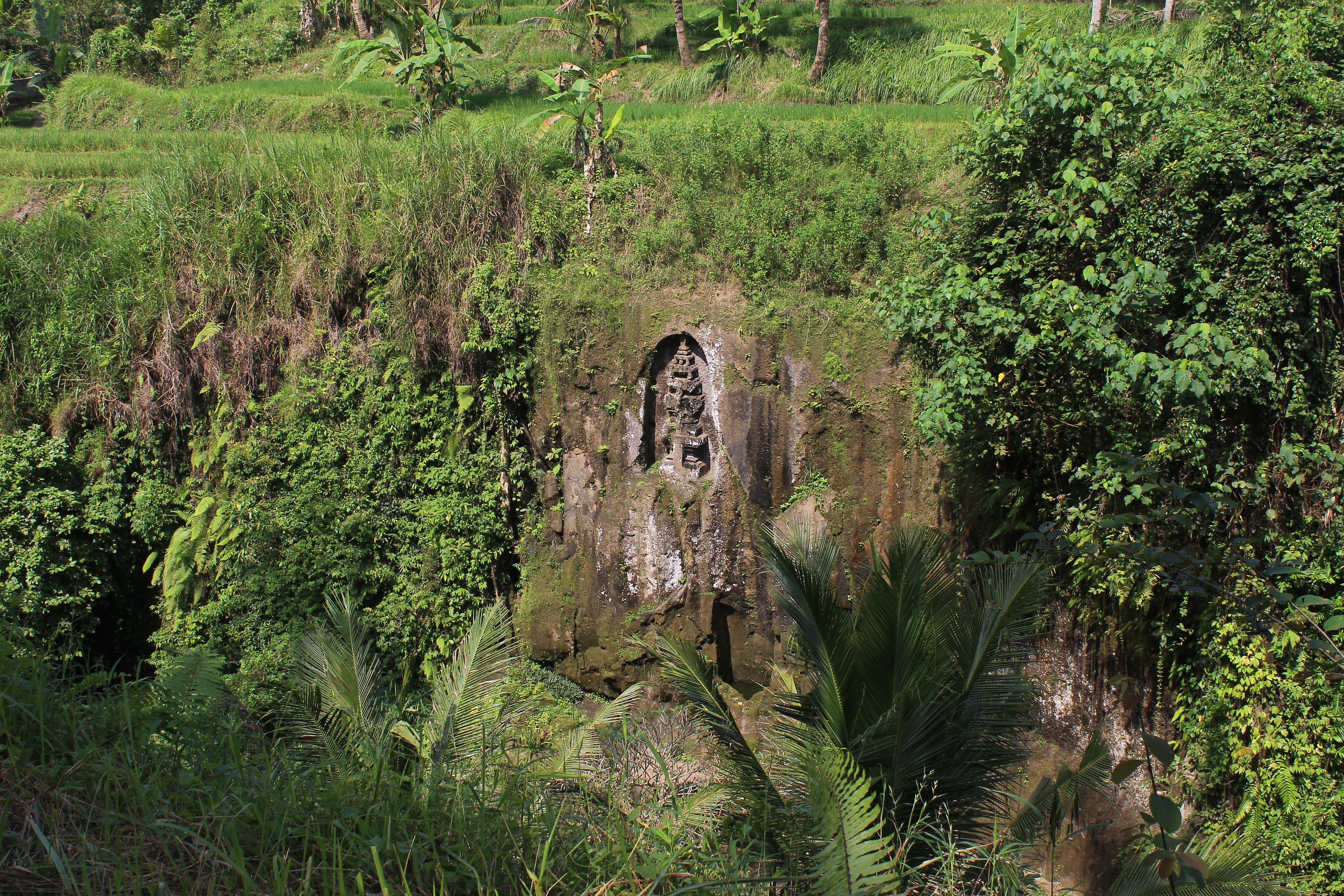 Small rock-cut shrine in the river ravine, with rice paddies along the upper bank