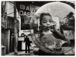 A child blows a huge bubble while a sailor stands in the far background