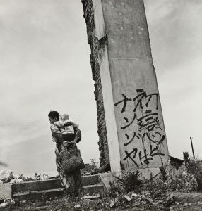 Black and white photo of a mother and child standing by a vertical structure with japanese writing on it