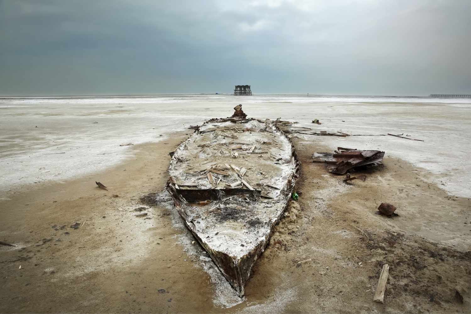 Photograph showing the frame of a rotting boat embedded in the sand of a dry lake bed.
