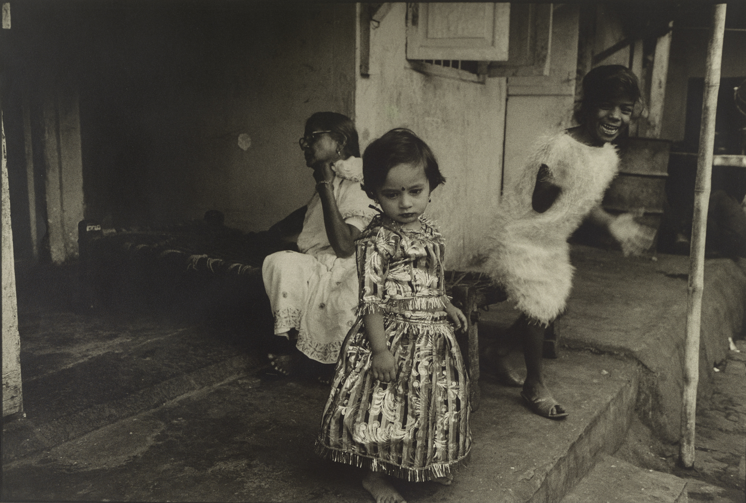 In a black-and-white photograph, one woman and two girls are gathered on the stoop outside a building. On the left, the woman in glasses wears white traditional dress and sits calmly, casting her gaze outside the frame. On the right, one little girl wearing a white feathery dress grins excitedly as she  rises from her seat, her hands blurred with motion. Front and center, the youngest little girl wears a striped dress and a bindi and looks downward with a serene expression.