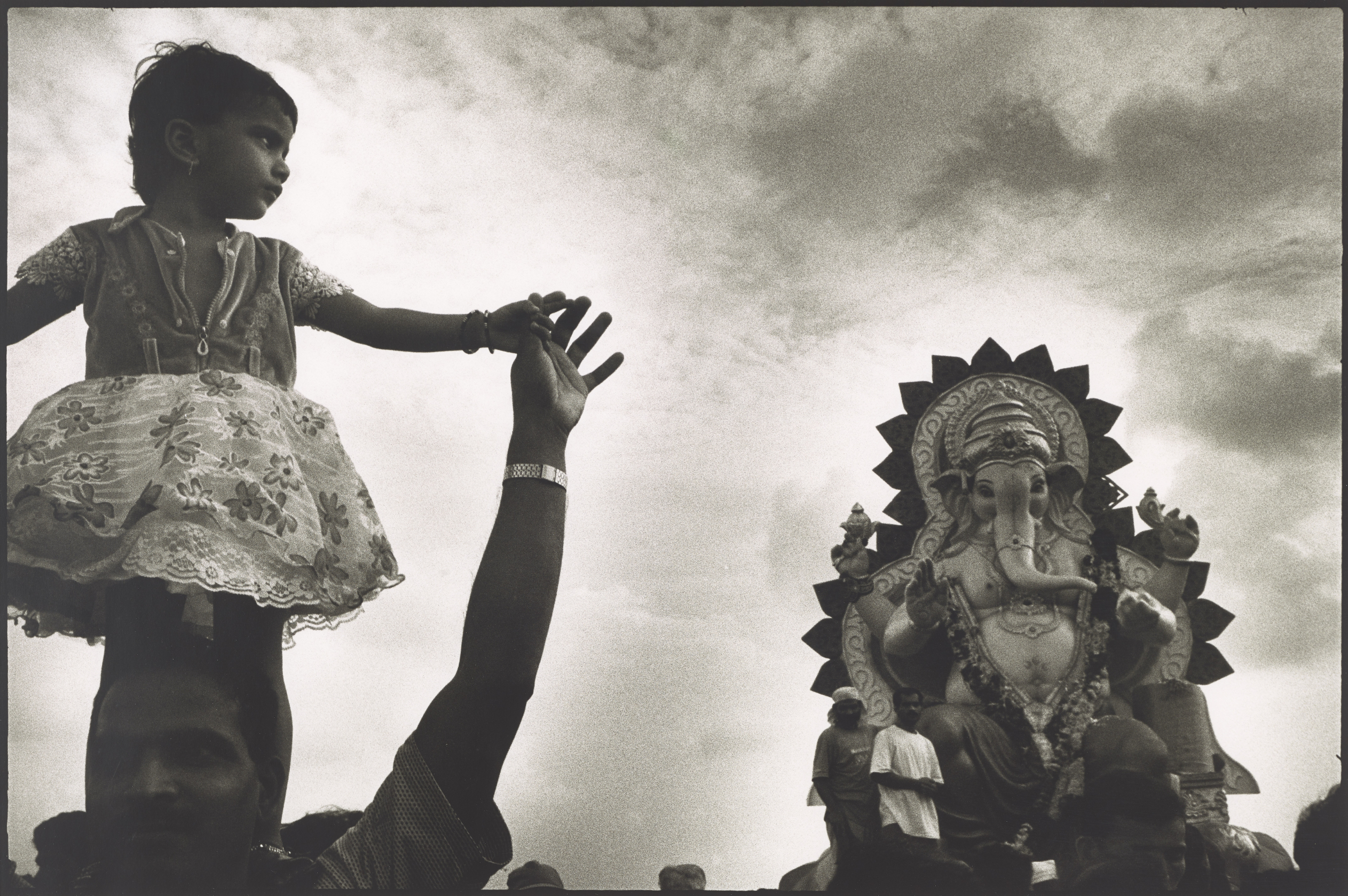 On the left in a black-and-white photograph, a young girl with short hair and a flowered dress stands on a man’s shoulders and holds his upstretched hand. She looks over her left shoulder at a large statue of the elephant-headed Hindu god Ganesh enthroned on a pedestal and adorned with garlands. The statue is at least twice as tall as the men beside it.